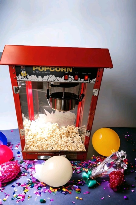 Popcornmaschine, die in einem gemütlichen Raum steht. Sie besteht aus einem metallenen Gehäuse, einem transparenten Glasbehälter und einer rot-weißen Markierung. Popcorn sprudelt aus einem Schlitz oben heraus, während der Boden mit leeren Popcorn-Tüten dekoriert ist